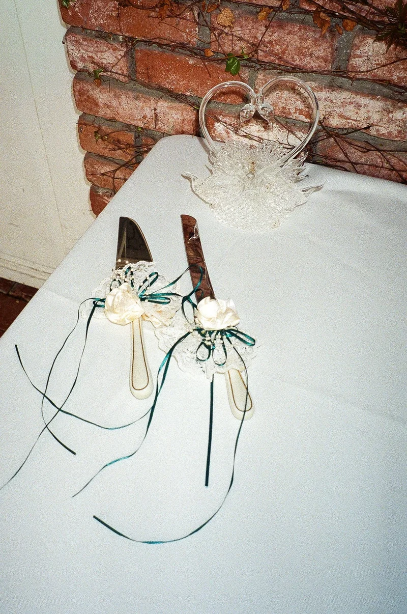 Two ribbon-tied cake knives on a white tablecloth beside a glass swan, brick wall behind