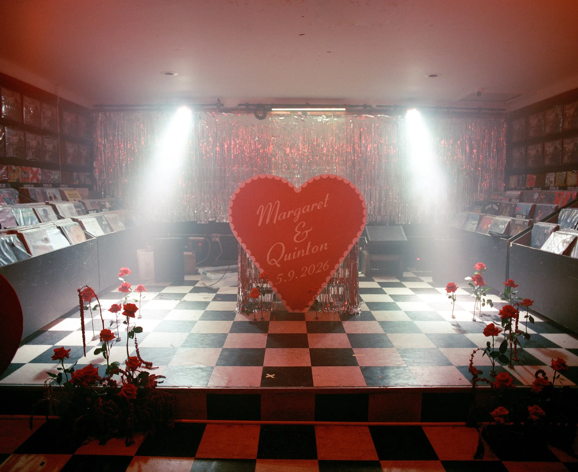 A record shop set for a wedding: checkerboard dance floor, silver tinsel curtain, and a large red heart sign reading Margaret & Quinton, lined with red roses