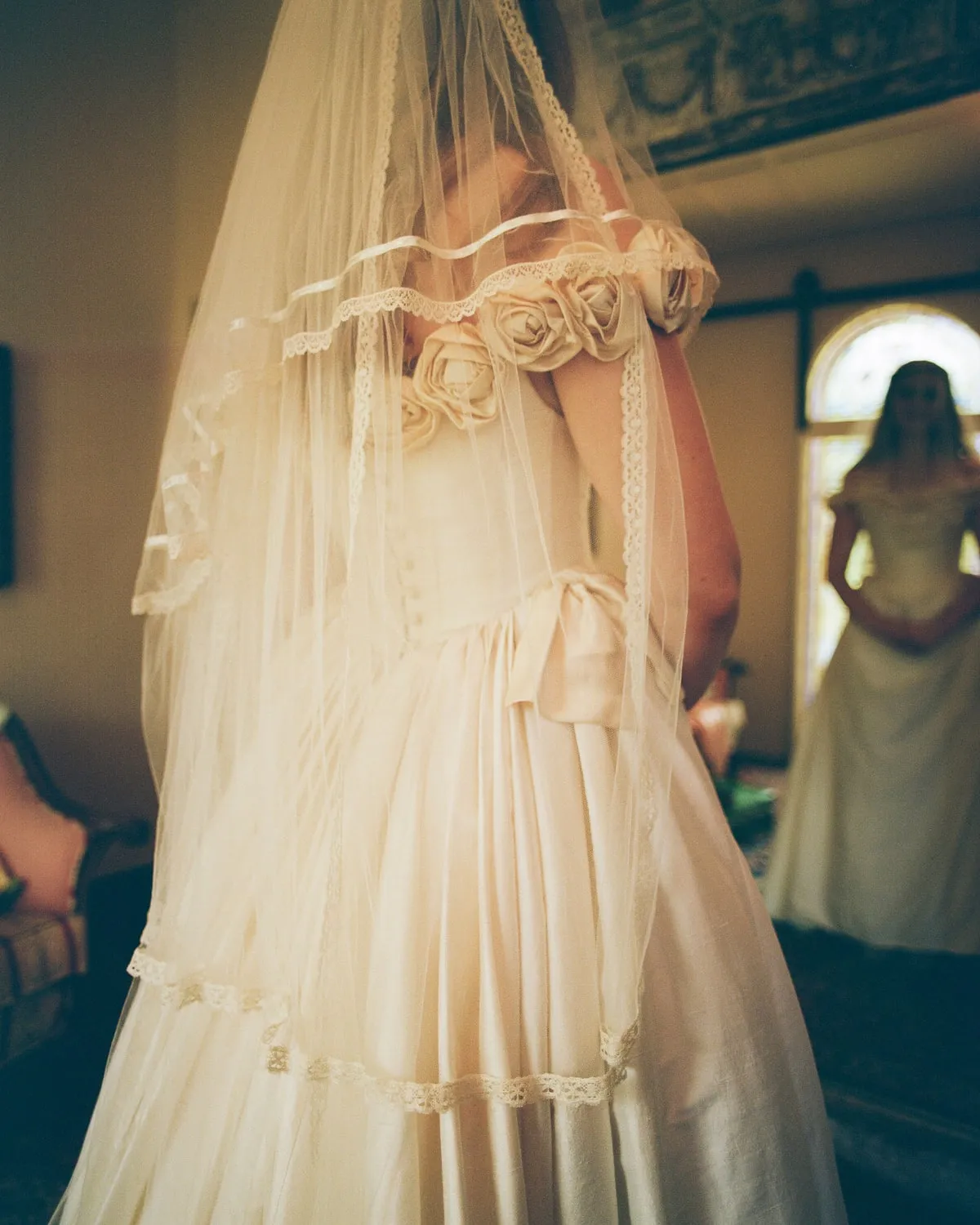 Film scan of a bride in a lace-trimmed veil and gown, a second bride reflected in an arched window behind her