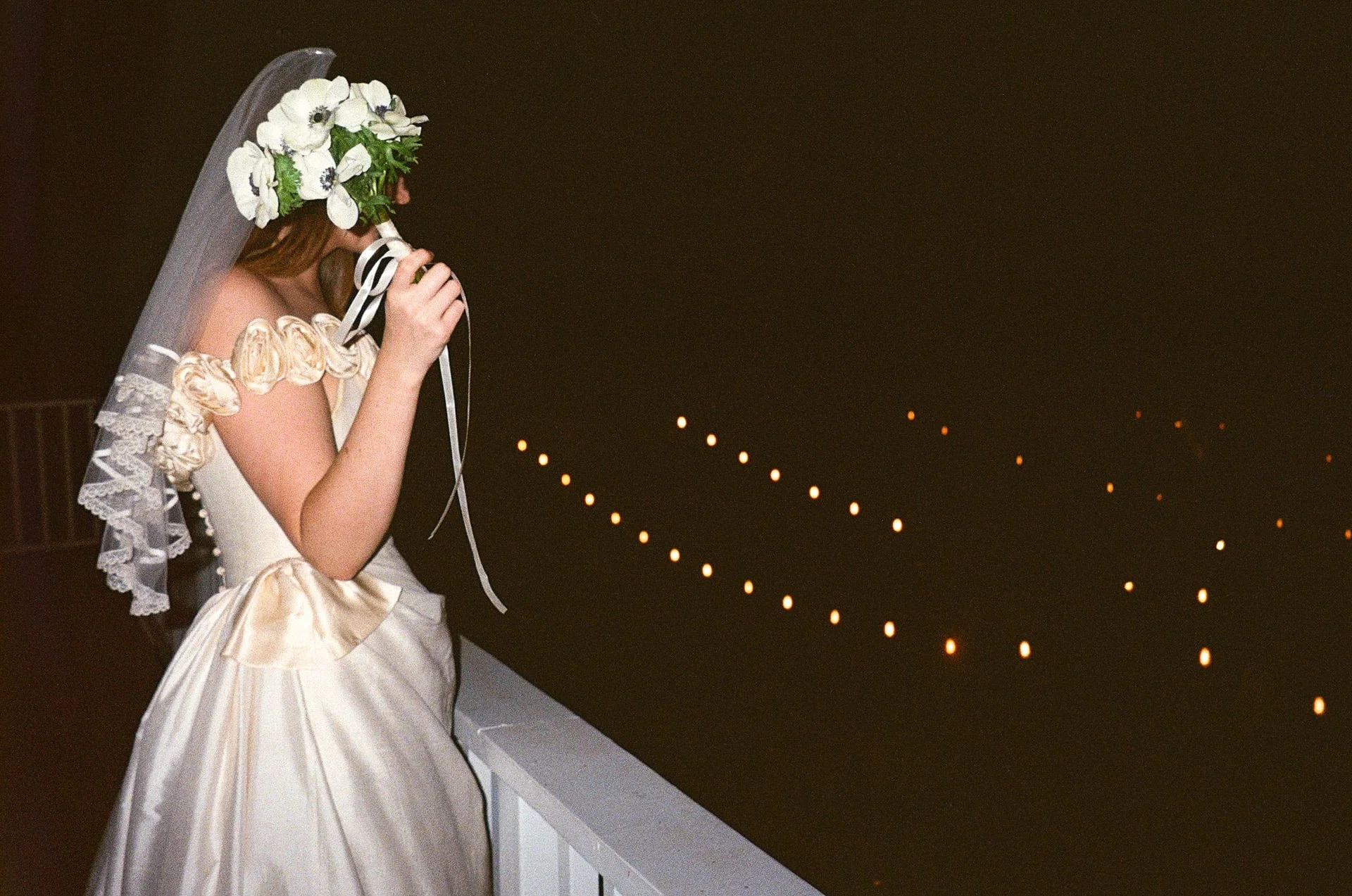 Bride on a balcony at night lifting her bouquet of white anemones to her face, string lights glowing behind her