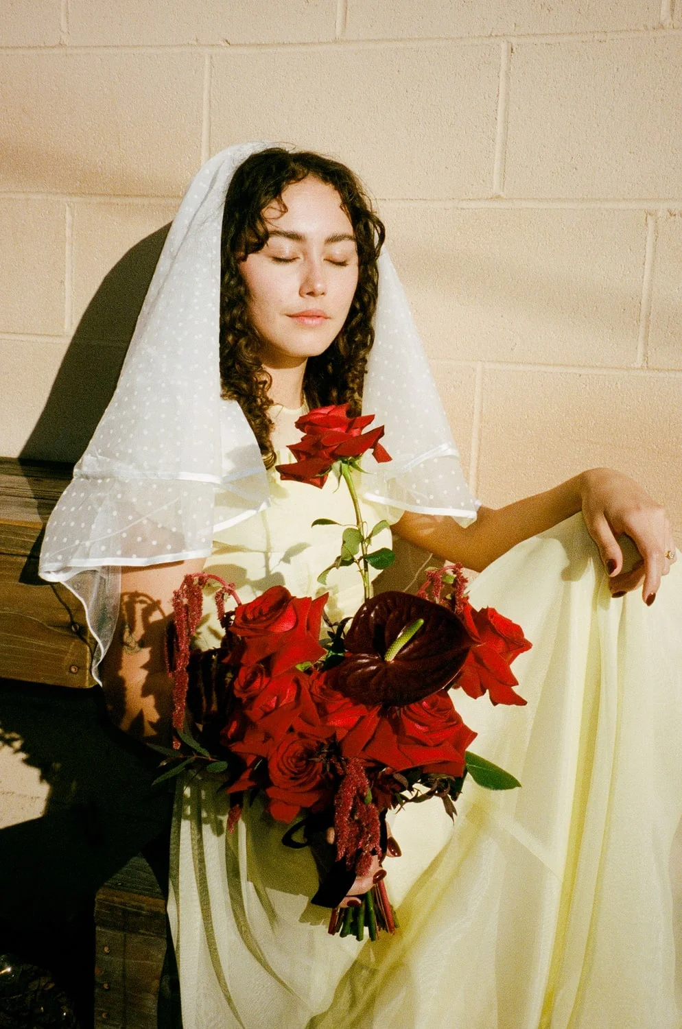 Bride in a dotted veil and pale yellow dress against a cinderblock wall, holding deep red roses and anthurium in hard sun