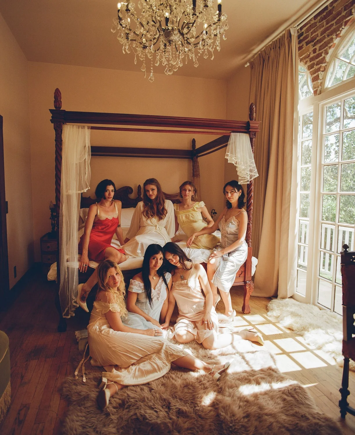 Seven bridesmaids in slip dresses gathered on and around a carved four-poster bed beneath a chandelier