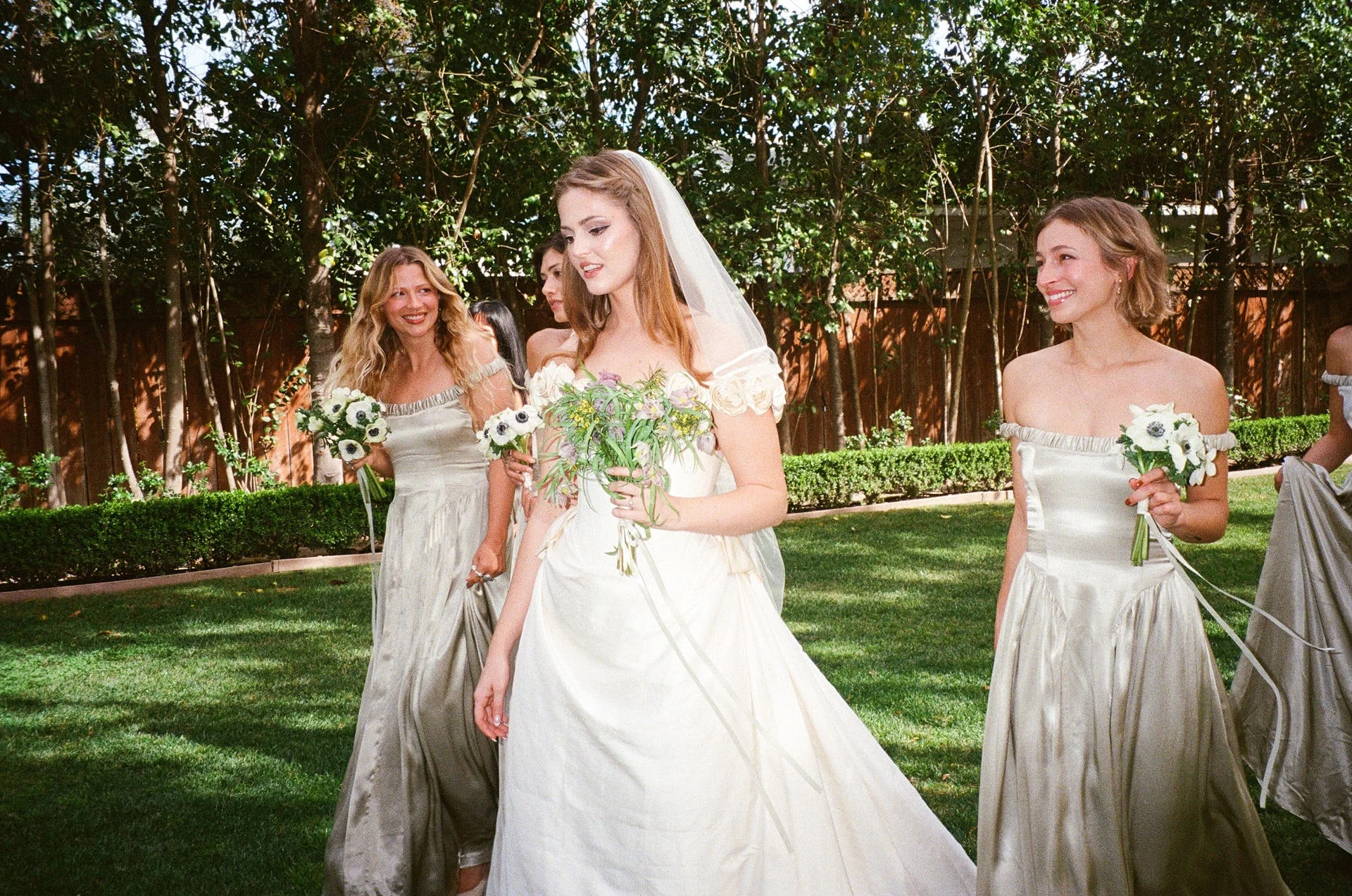 Bride walking across a lawn with her bridesmaids in sage slip dresses, anemone bouquets in hand