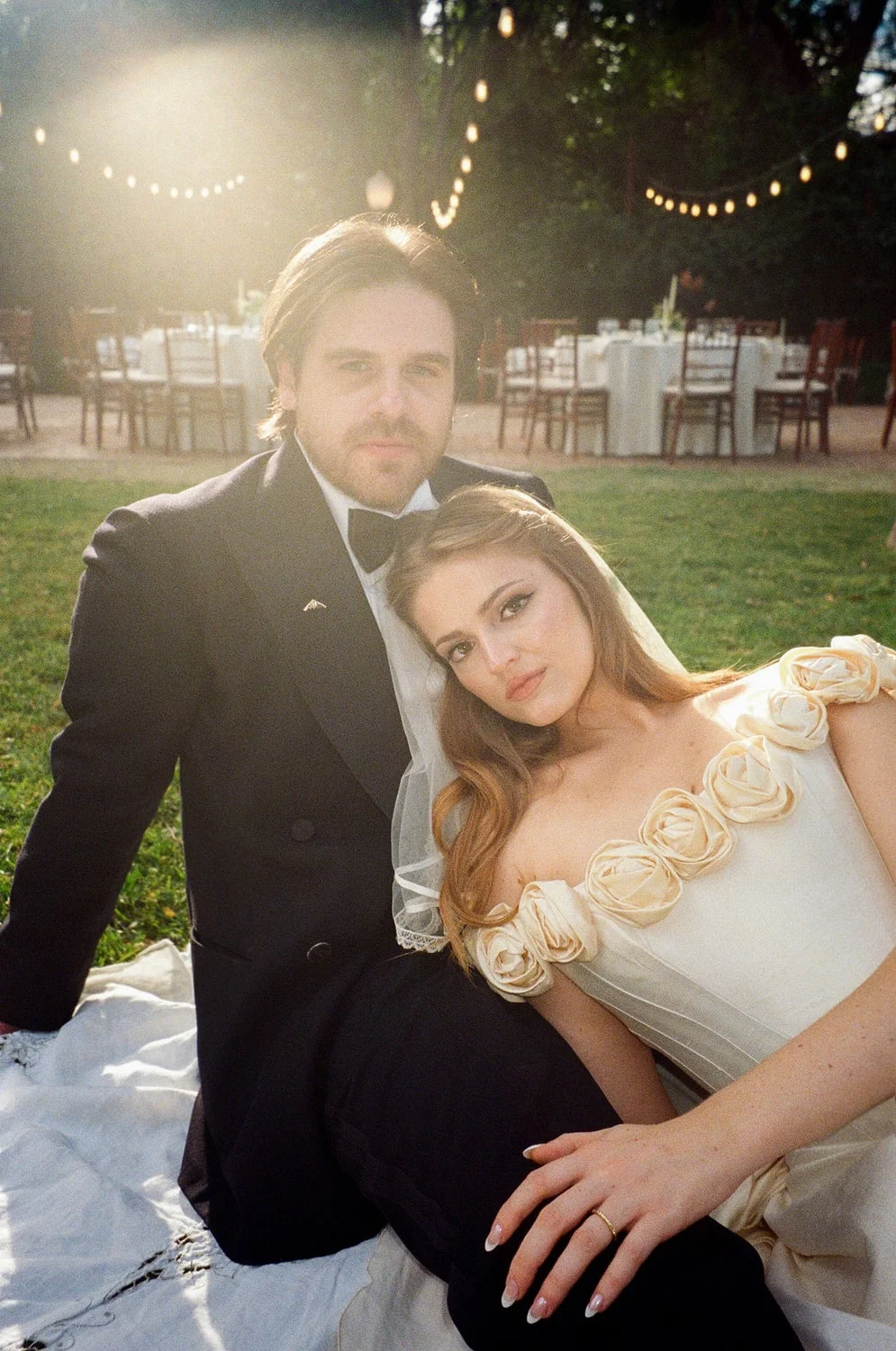 Couple resting together on a blanket on the grass at golden hour, set tables and string lights behind them