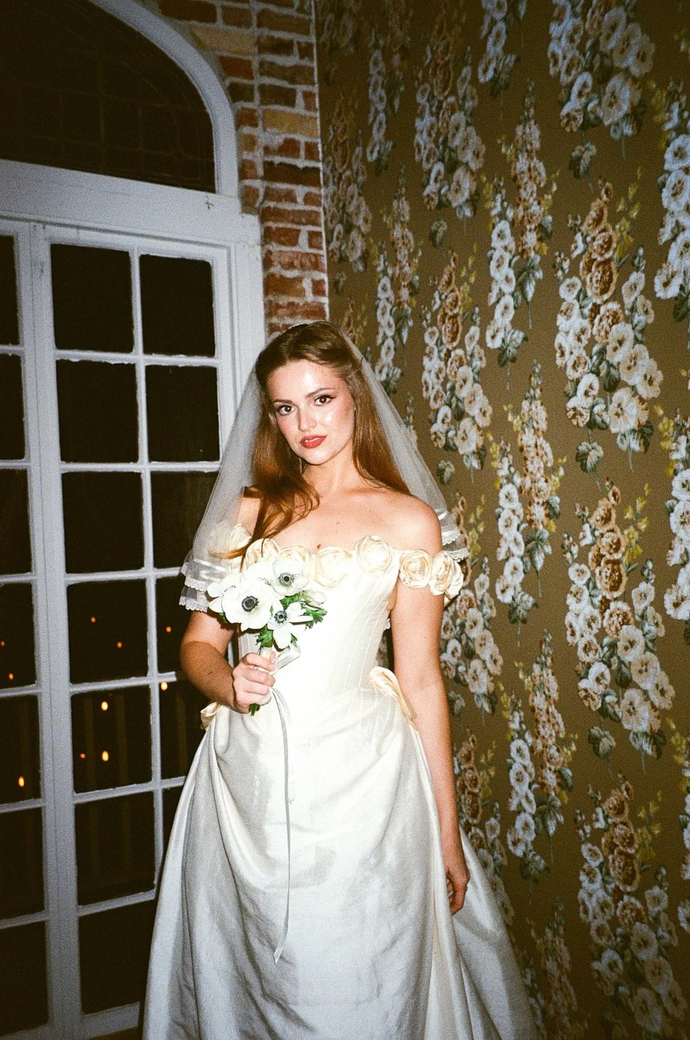 Bride in an ivory gown and veil holding white anemones, lit by flash against floral wallpaper and bare brick