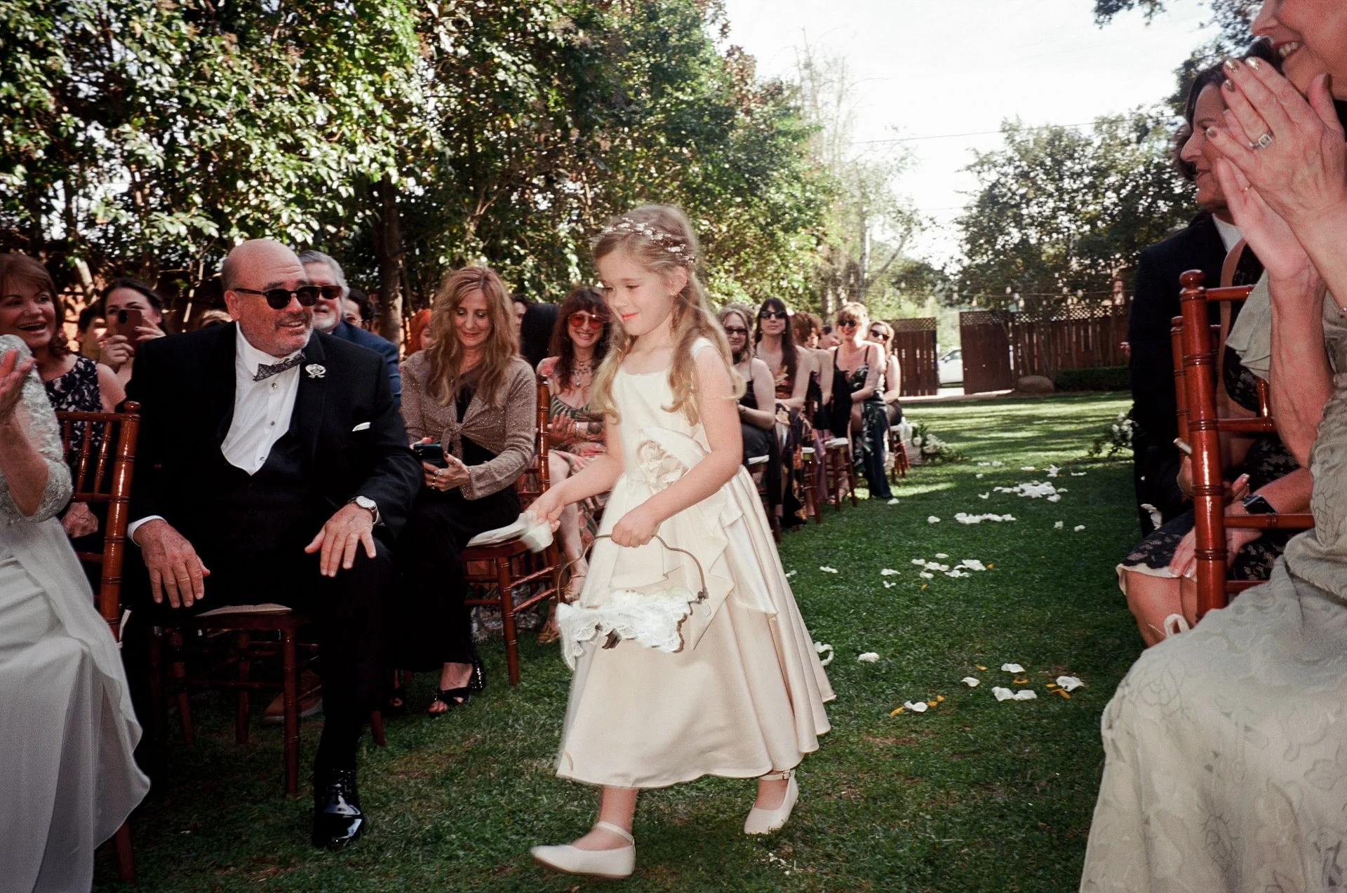 Flower girl walking a grass aisle scattered with petals as seated guests clap