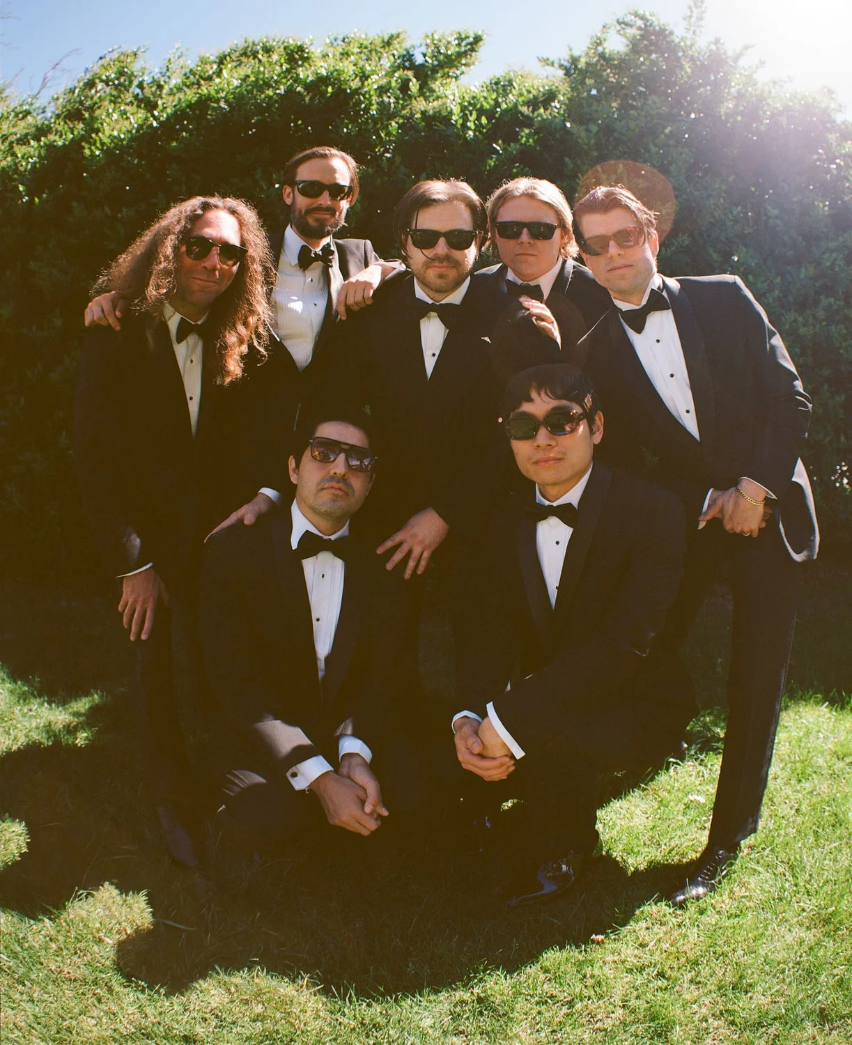Seven groomsmen in tuxedos and sunglasses grouped together on a sunlit lawn