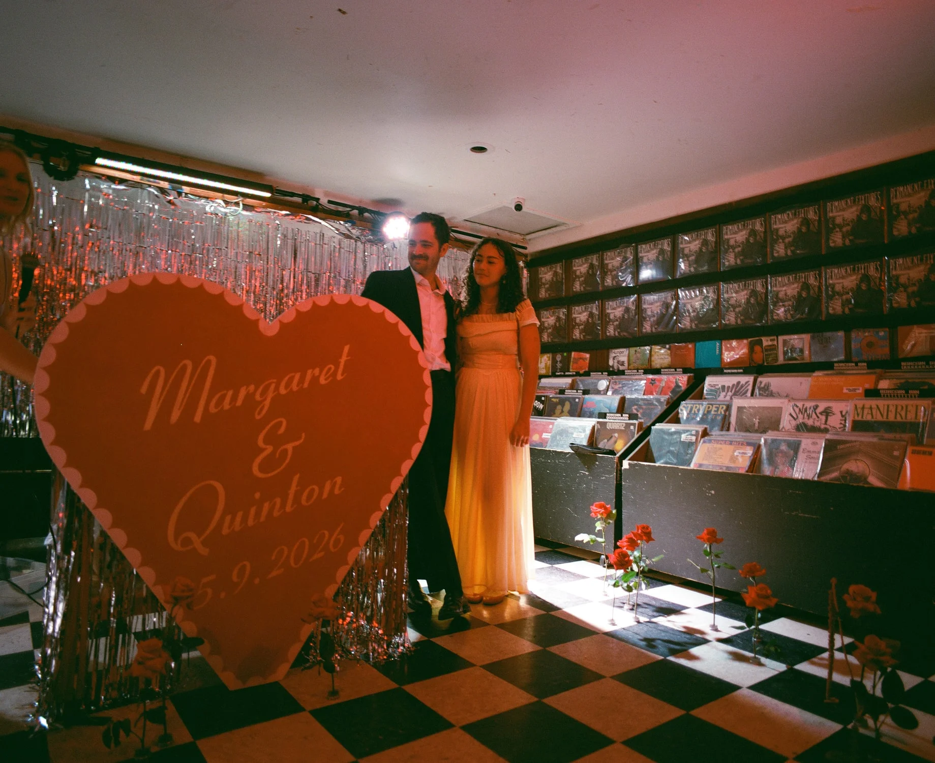 Couple standing beside a tall red heart sign in a record shop, tinsel curtain and record bins around them