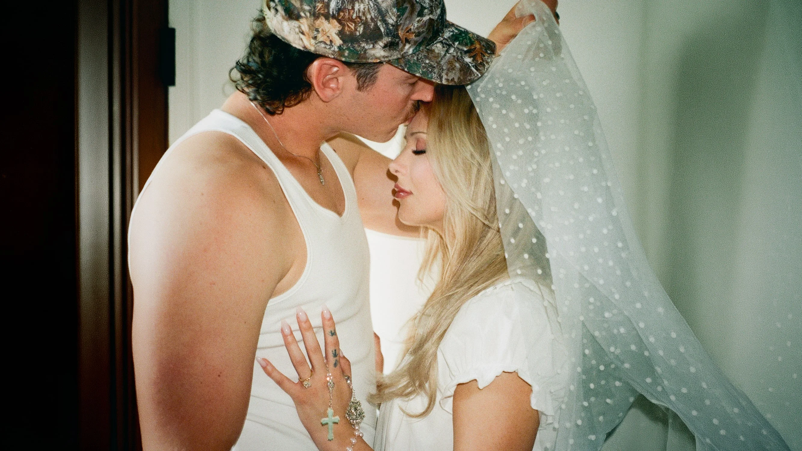 Groom in a camo cap kissing his bride on the forehead beneath a dotted veil