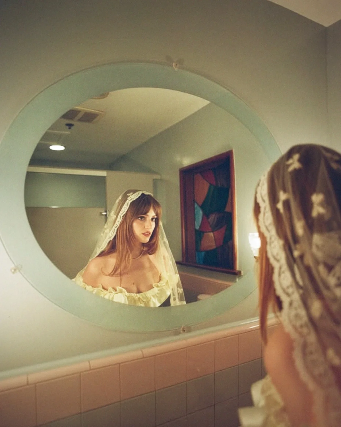 Bride in a lace veil reflected in a round mirror in a pink tiled bathroom
