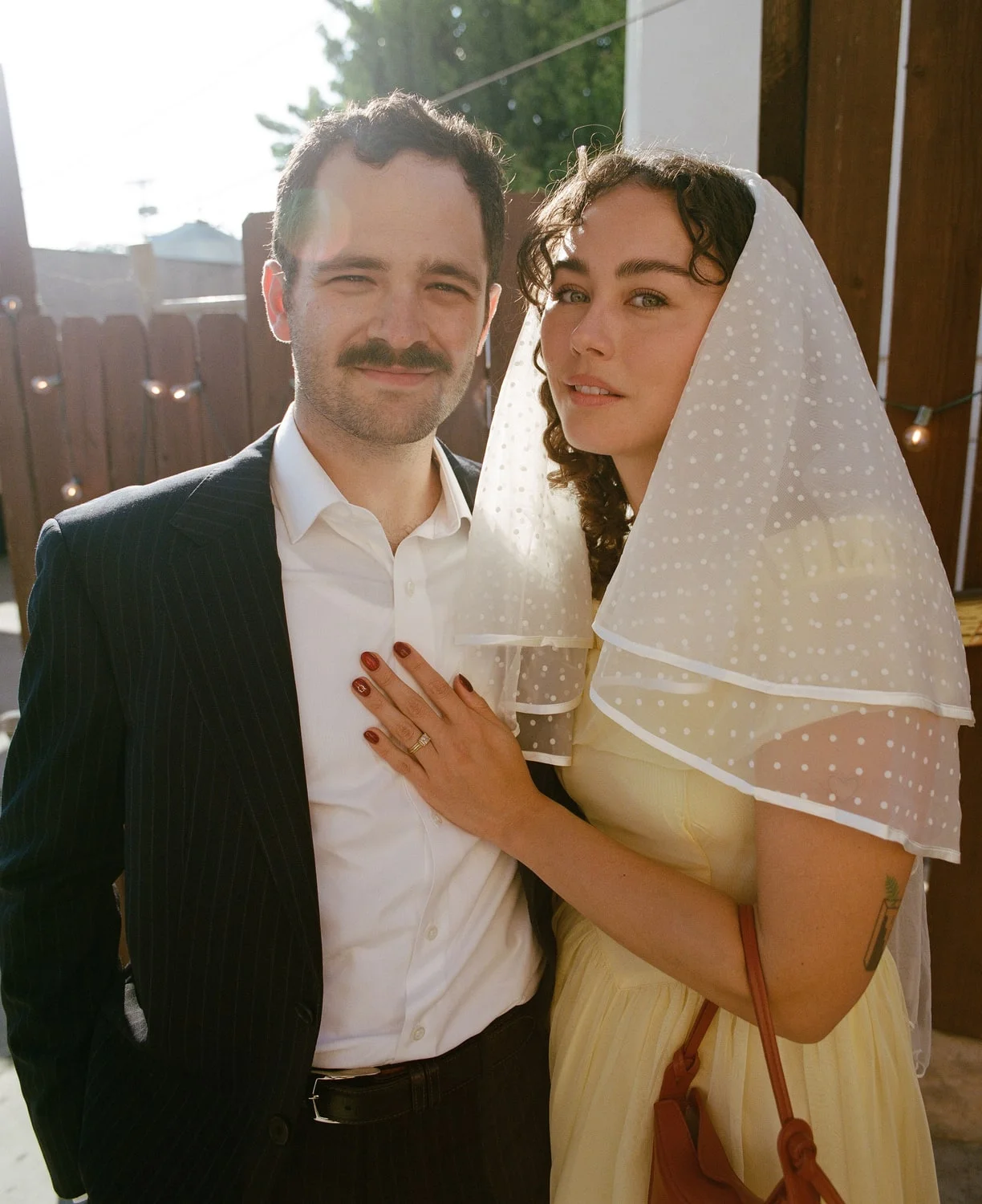 Couple close together in late sun, the groom in a pinstripe suit and the bride in a yellow dress and dotted veil