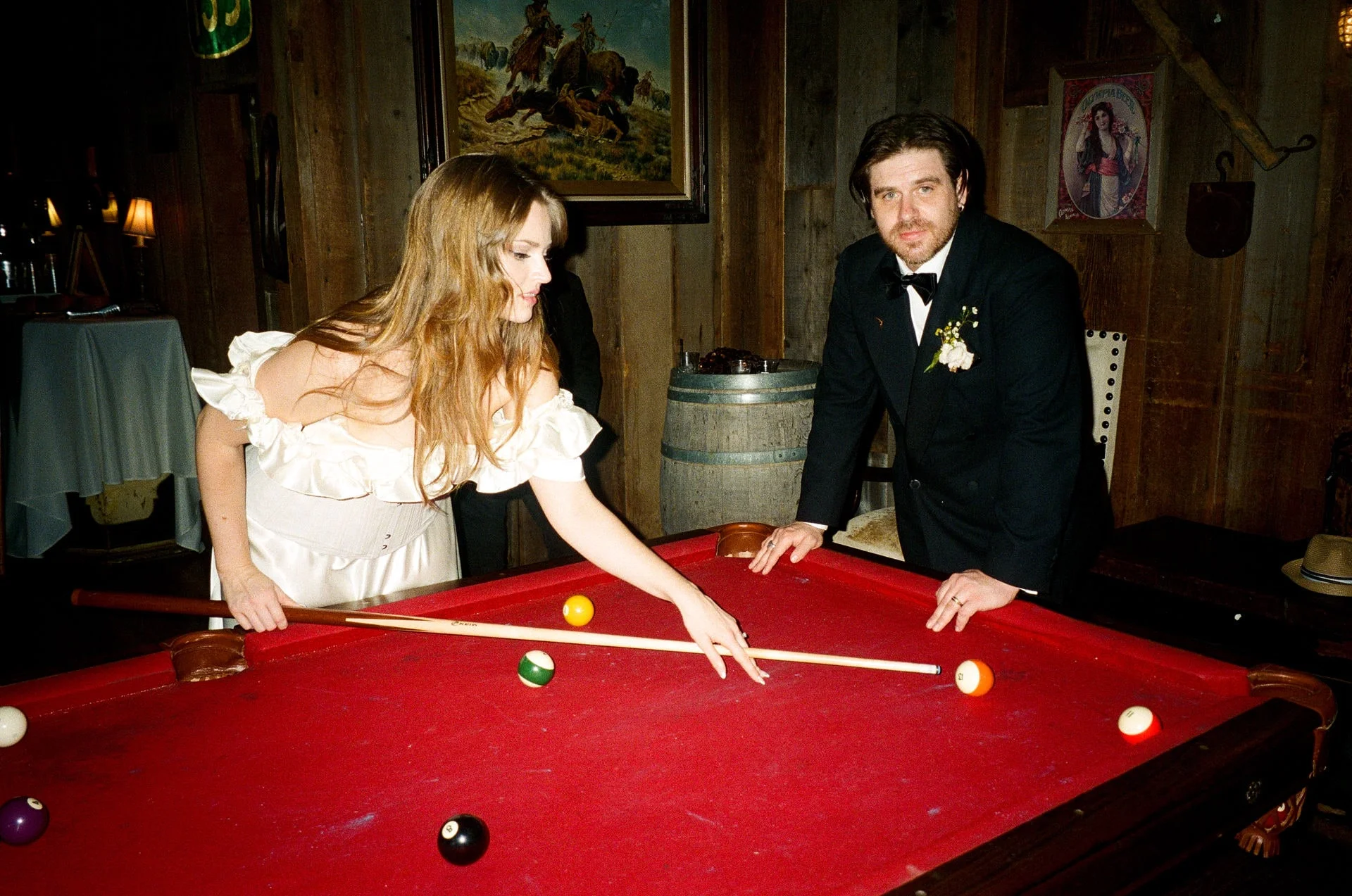 Bride in a ruffled white dress leaning across a red-felt pool table to line up a shot while her groom watches from the far rail
