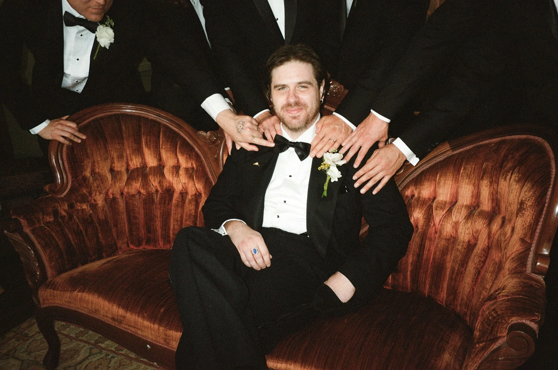 Groom seated on a rust velvet settee, his groomsmen standing behind him with hands on his shoulders