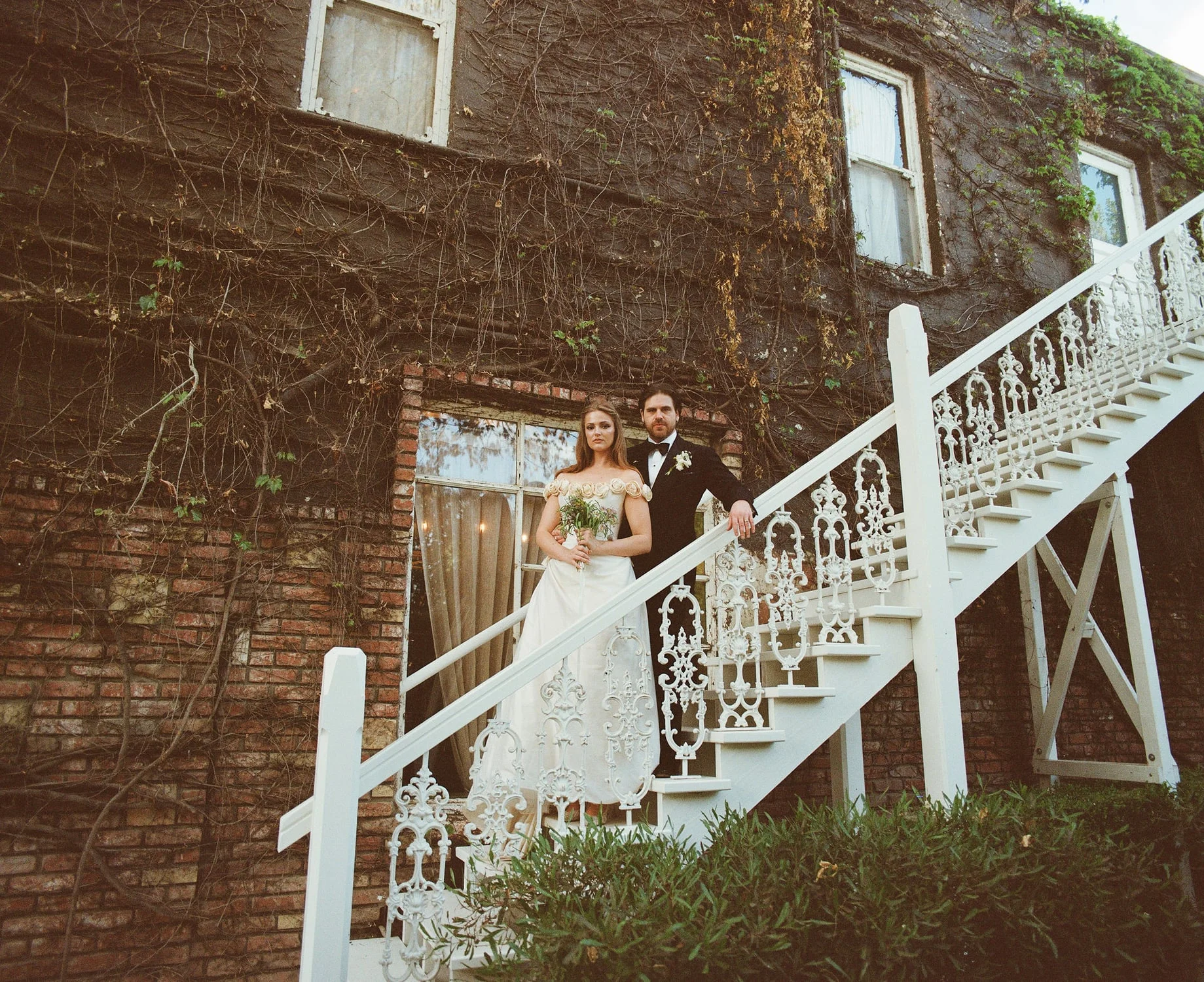 Couple standing on a white ironwork staircase against an ivy-covered brick building