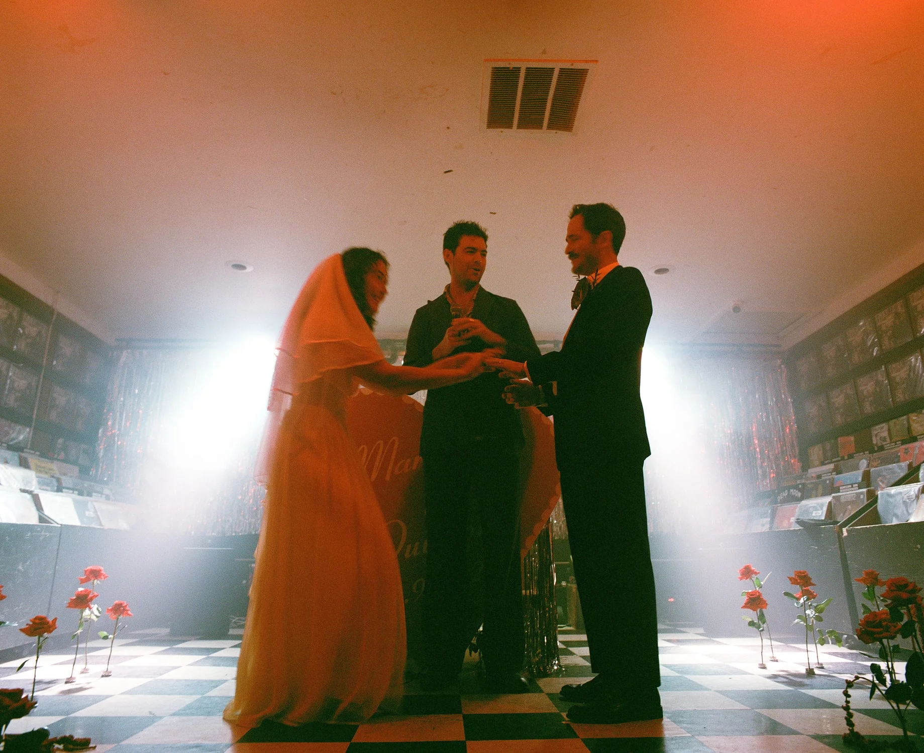 Couple exchanging rings in front of their officiant on a checkerboard floor in a record shop, backlit by haze